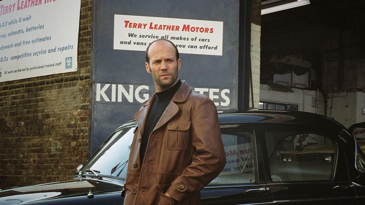 Jason Statham filmy. Mężczyzna w brązowej skórzanej kurtce stoi przed czarnym samochodem, w tle widać szyld "Terry Leather Motors".