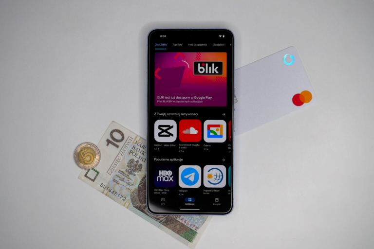 Smartfon wyświetlający ekran Google Play z aplikacją BLIK, obok leżą polskie pieniądze - banknot dziesięciozłotowy i moneta pięciozłotowa oraz biała karta płatnicza.