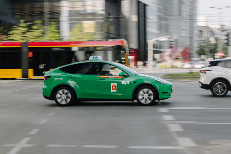 Zielona taksówka elektryczna Bolt marki Tesla jadąca po ulicy obok białego samochodu i żółto-czerwonego tramwaju na tle budynków miejskich.