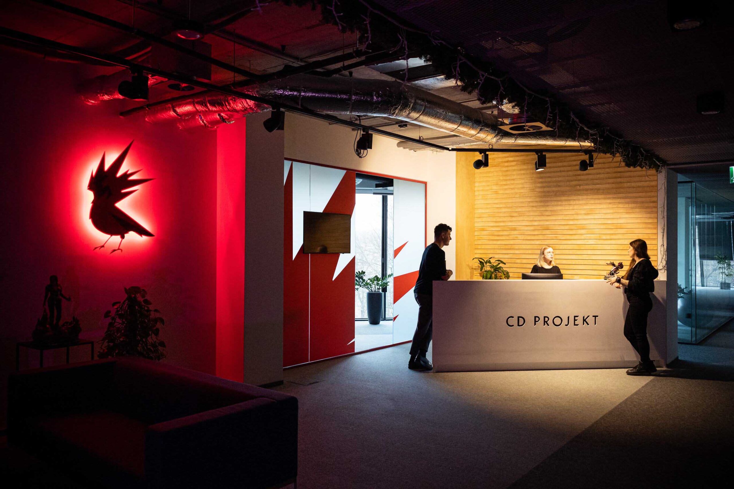 Recepcja CD Projekt z trzema osobami, oświetlonym logo czerwonego ptaka na ścianie po lewej stronie.