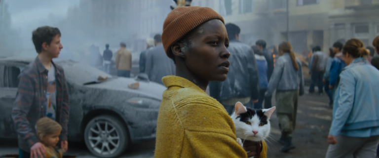 Kobieta w pomarańczowej czapce trzyma kota na tle zatłoczonej, zadymionej ulicy wypełnionej ludźmi i pokrytymi popiołem samochodami.