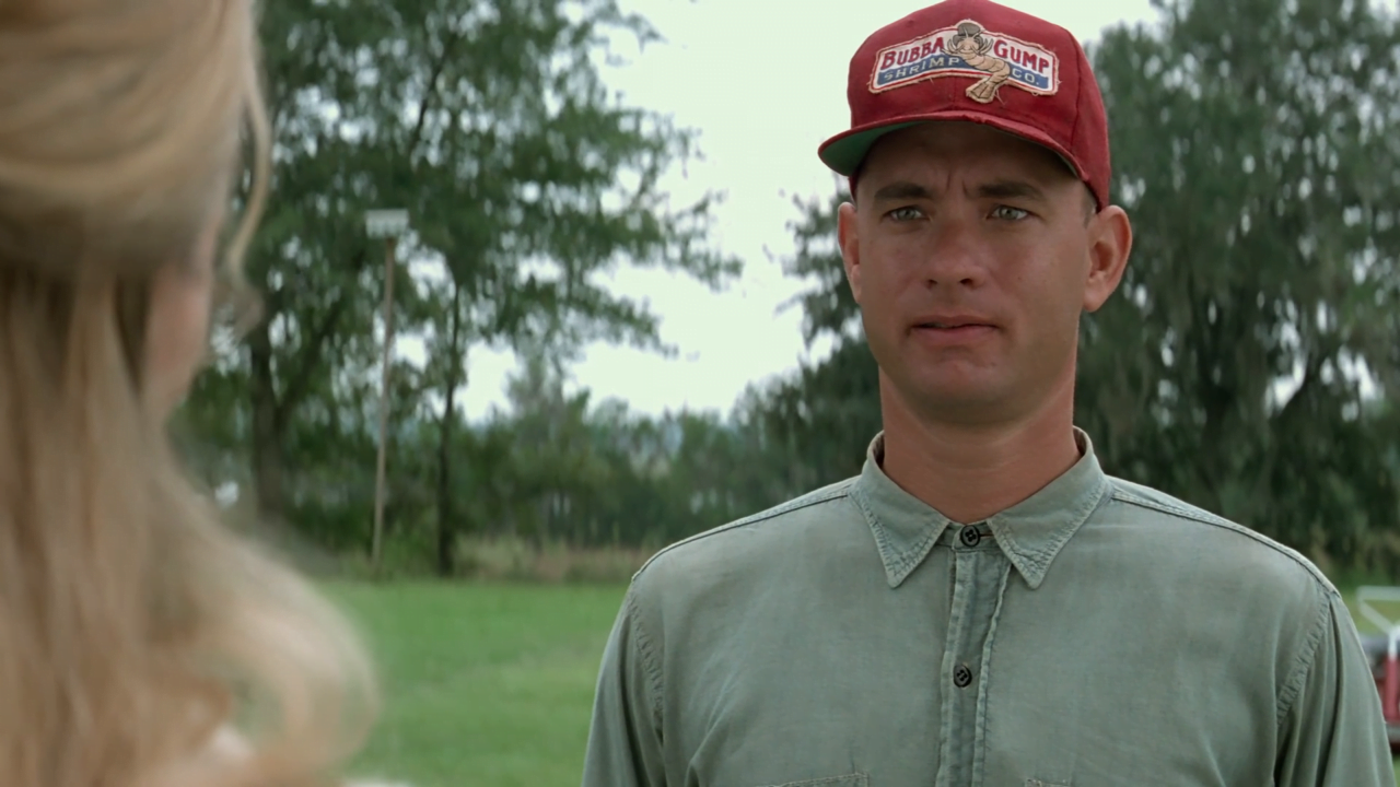 Tom Hanks filmy. Mężczyzna w czerwonej czapce "Bubba Gump Shrimp Co." i szarej koszuli stoi na tle drzew.