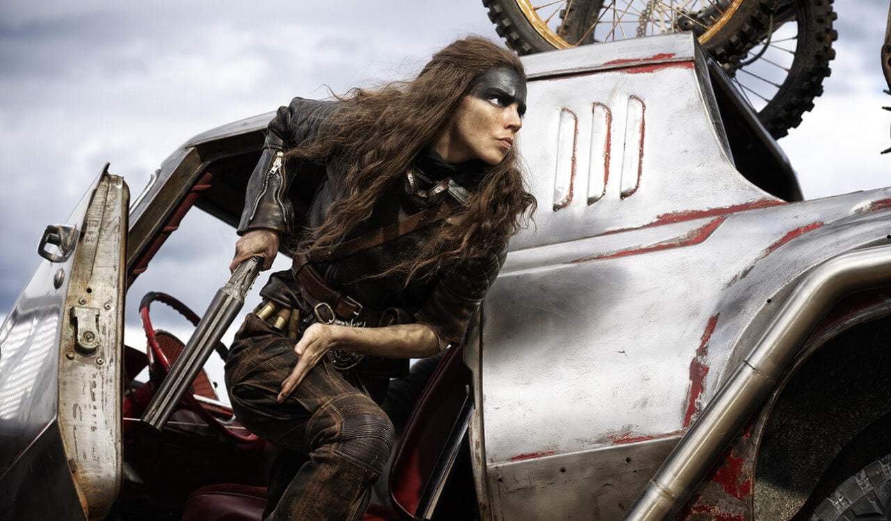 Kadr z filmu Furiosa. Osoba z długimi włosami, w skórzanym stroju, wychodząca z metalowego samochodu z dwururką w ręku.