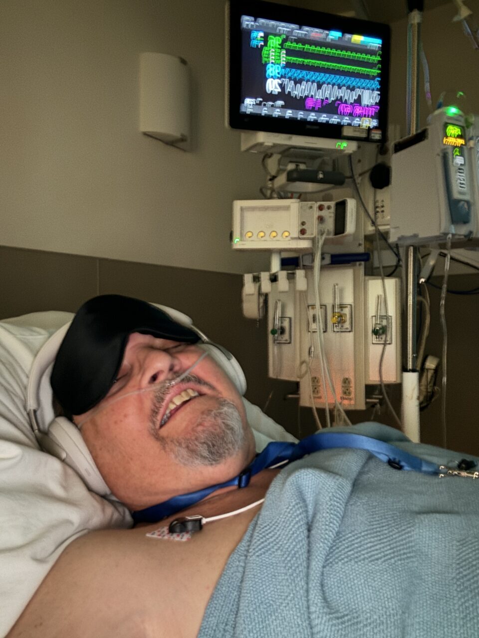 Mężczyzna leżący w szpitalnym łóżku z założoną maską tlenową i słuchawkami, obok urządzenia z wyświetlaczem monitorującym parametry życiowe.