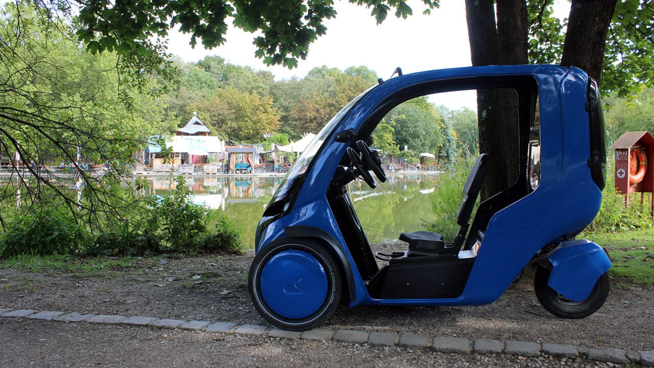 Mały, niebieski, nowoczesny pojazd elektryczny zaparkowany na bruku przy parku z widokiem na staw i budynki w tle.