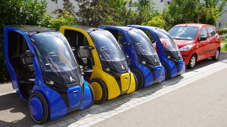 Małe elektryczne pojazdy trójkołowe w kolorze niebieskim i żółtym zaparkowane obok większego czerwonego samochodu osobowego.