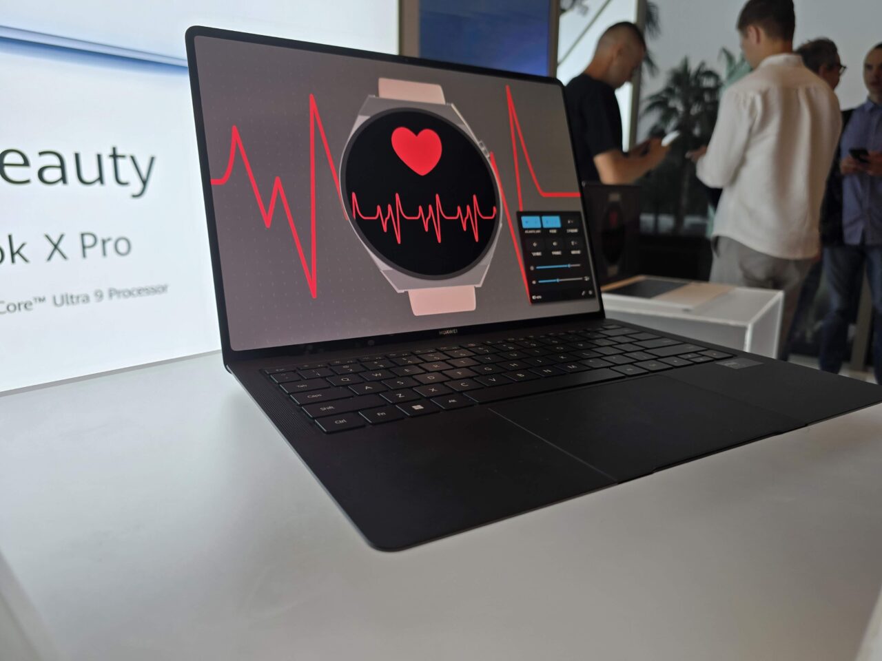 Laptop Huawei MateBook X Pro 2024 em exibição com uma tela mostrando smartwatch e gráficos de batimentos cardíacos, com figuras desfocadas de pessoas ao fundo.