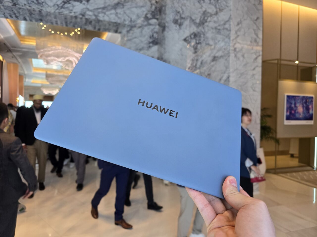 Um portátil Huawei MateBook X Pro 2024 azul segurado numa mão contra o fundo de figuras desfocadas num interior elegante.