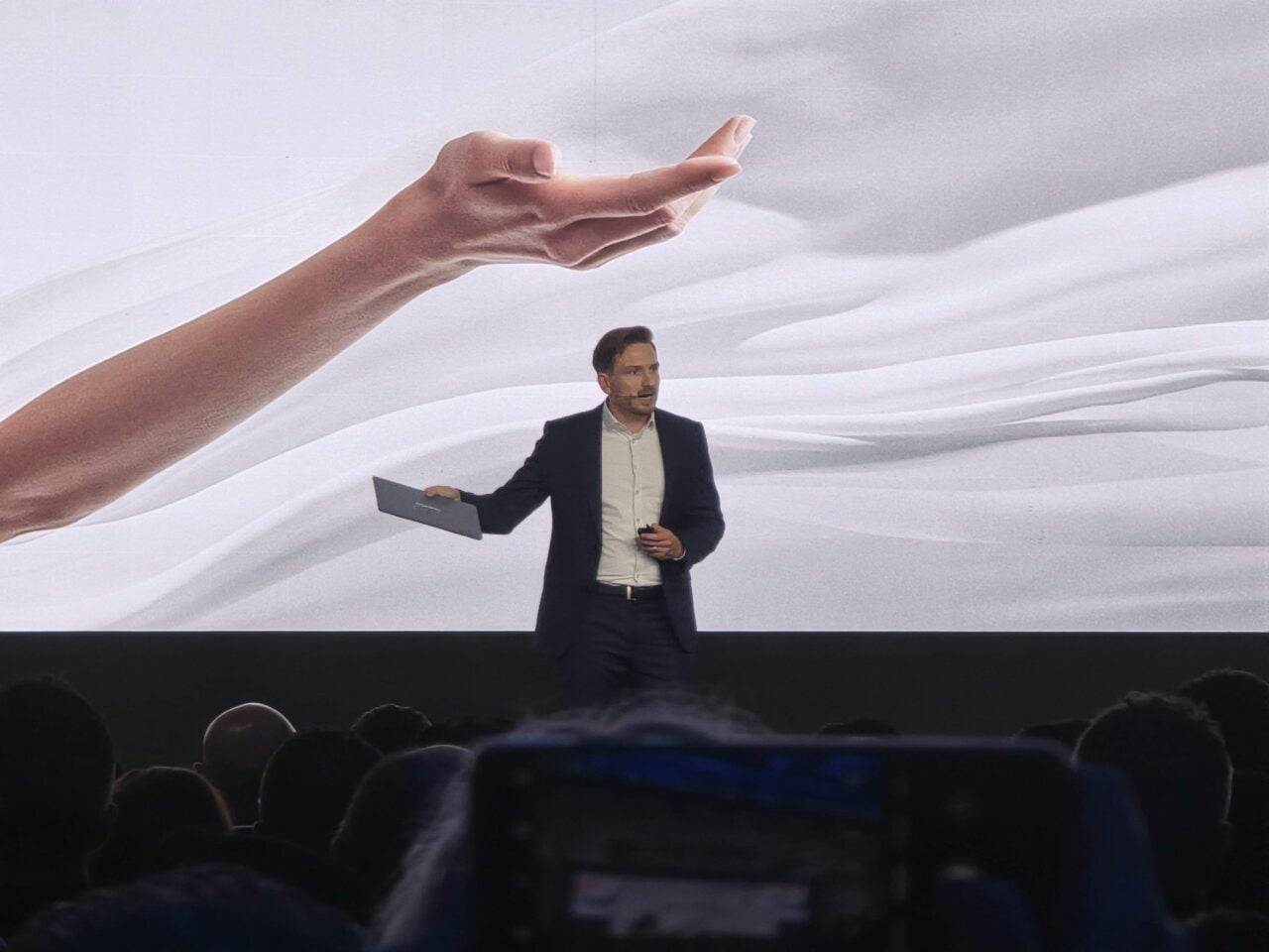 Mężczyzna prezentujący Huawei MateBook X Pro 2024 przed dużym ekranem z grafiką dłoni, na tle zgromadzonej publiczności i kamery filmującej.