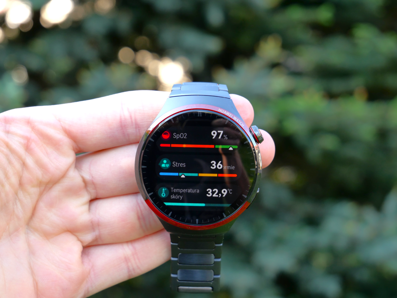 Smartwatch trzymany w dłoni, wyświetlający dane zdrowotne: poziom SpO2 97%, stres 36, temperatura skóry 32,9°C.