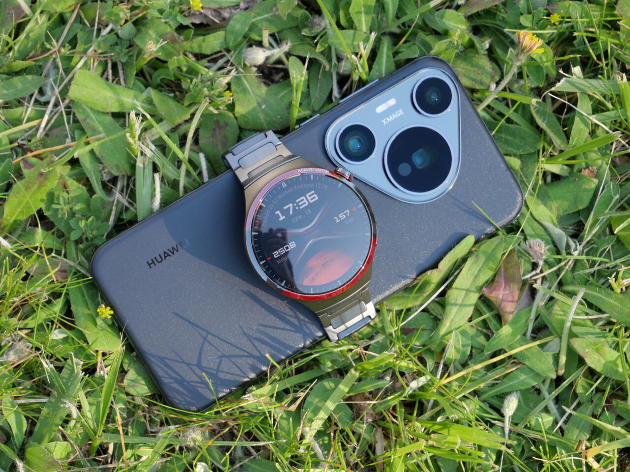 Smartfon Huawei oraz smartwatch z wyświetlaczem pokazującym godzinę 17:36, leżące na trawie.