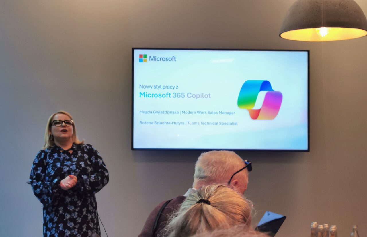 Microsoft Copilot em polonês.  Um palestrante ao lado de uma apresentação em uma tela grande com o logotipo da Microsoft e informações sobre o Microsoft 365 Copilot durante uma reunião de negócios.