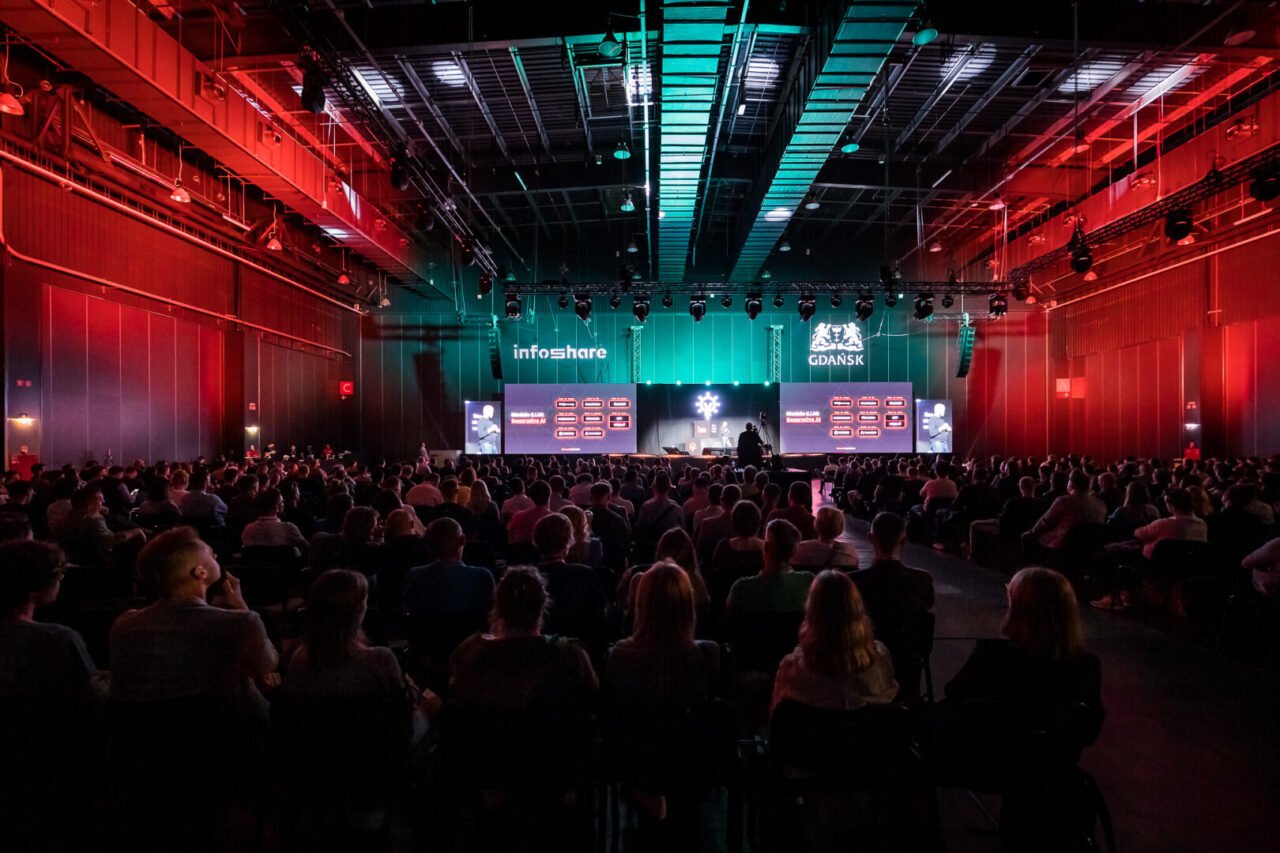 Público ouvindo uma apresentação na conferência de tecnologia InfoShare em uma sala de conferências iluminada em vermelho.