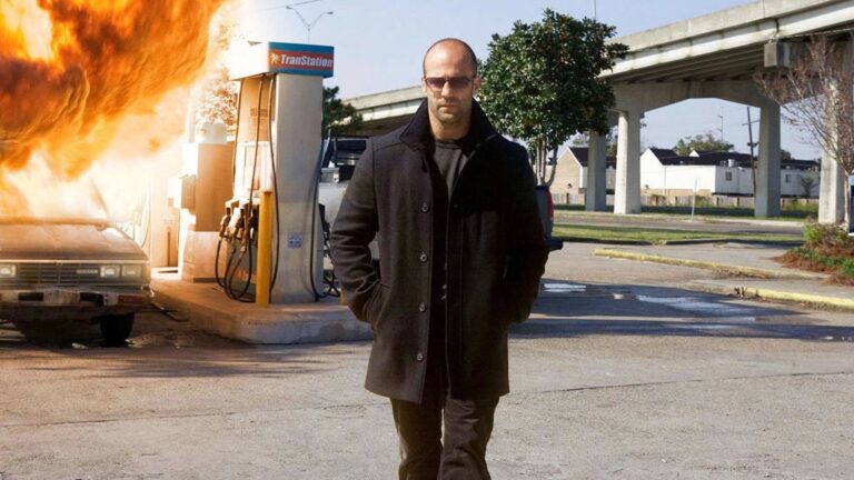 Mężczyzna w ciemnym płaszczu i okularach przeciwsłonecznych idzie spokojnie przed stacją benzynową, z eksplozją samochodu w tle.