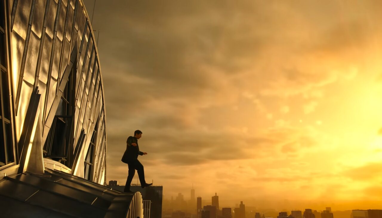 Kadr z filmu Megalopolis. Człowiek stojący na krawędzi budynku podczas wschodu lub zachodu słońca, z widokiem na panoramę miasta.