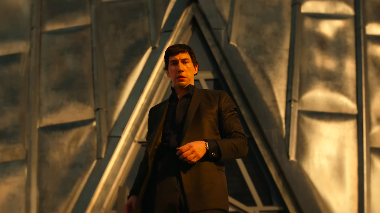 Kadr z filmu Megalopolis. Mężczyzna w garniturze patrzący w dół, stojący przed dużą, geometryczną strukturą z metalu lub betonu.