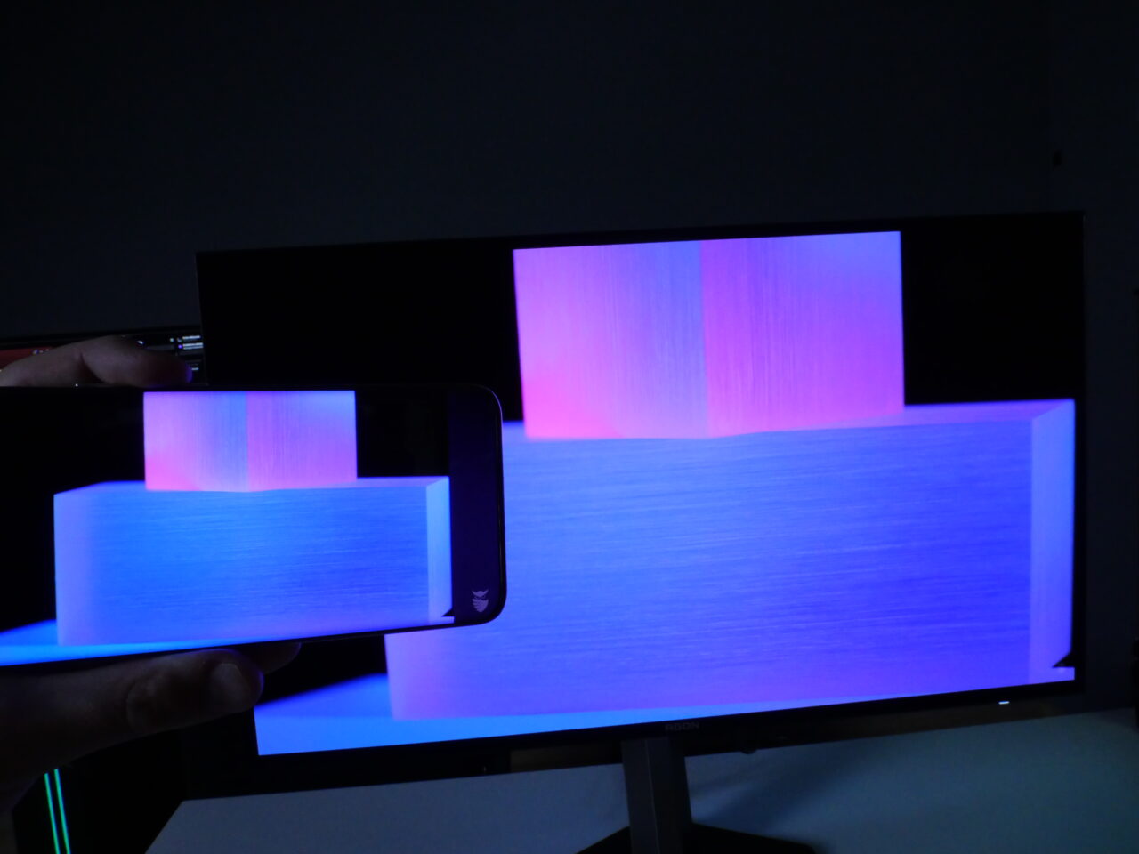 Ręka trzymająca smartfon, który wyświetla obraz przedstawiający różowe i niebieskie światła, na tle monitora komputerowego z tym samym obrazem.