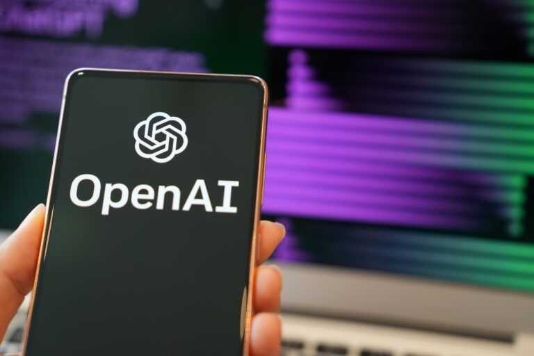 Smartfon trzymany w dłoni z logo OpenAI na ekranie na tle rozmytych kolorowych linii kodu.