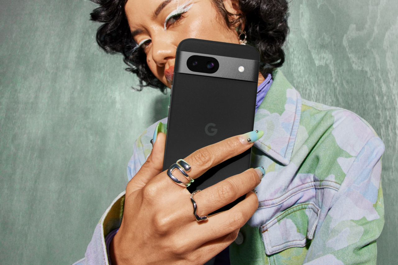 Cena Google Pixel 8  Kobieta robi selfie, trzymając smartfon z tyłu kamery w kierunku obiektywu. Ma na sobie kolorową kurtkę i stylowe pierścionki, a jej paznokcie są pomalowane na zielono z dekoracjami.