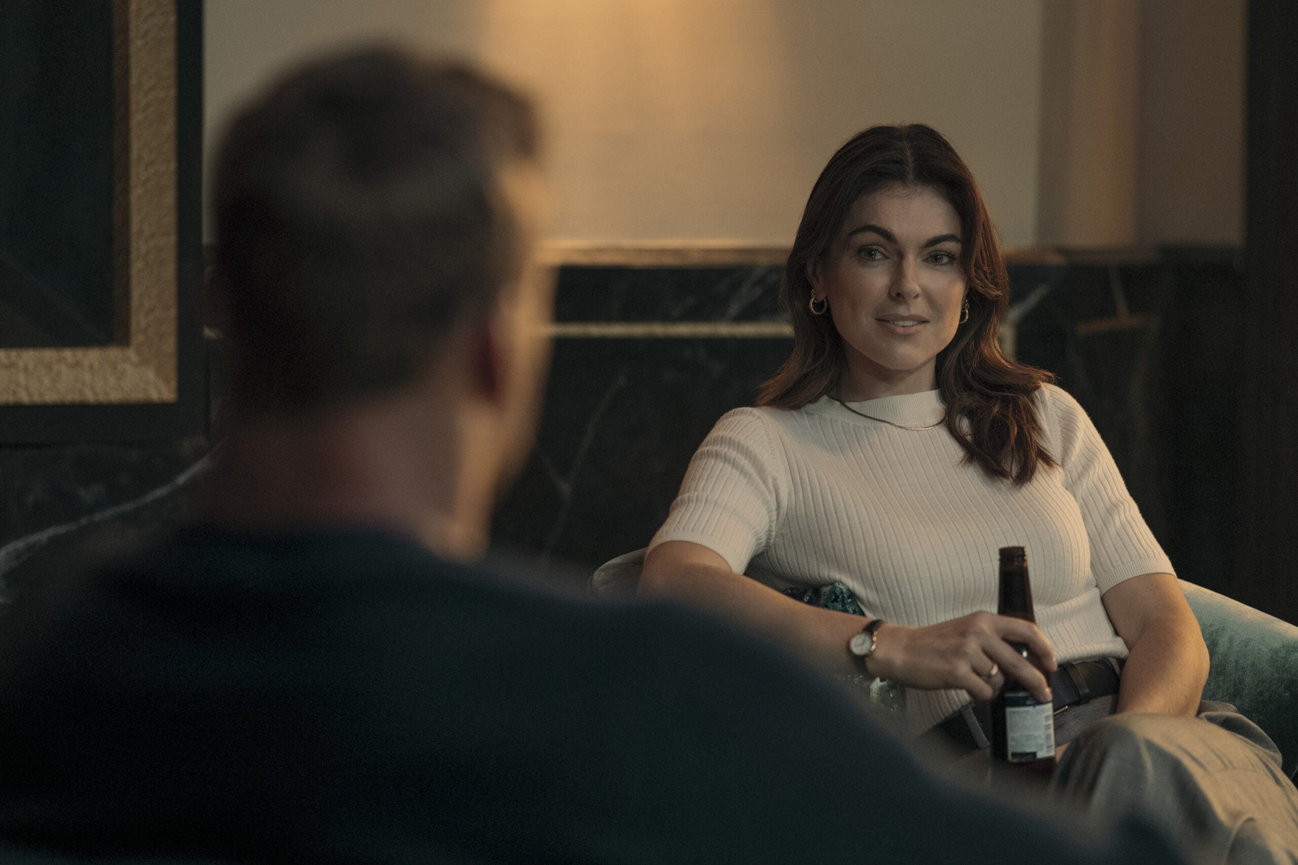 Kobieta siedząca na sofie trzymająca butelkę piwa, rozmawia z siedzącym naprzeciw mężczyzną.
