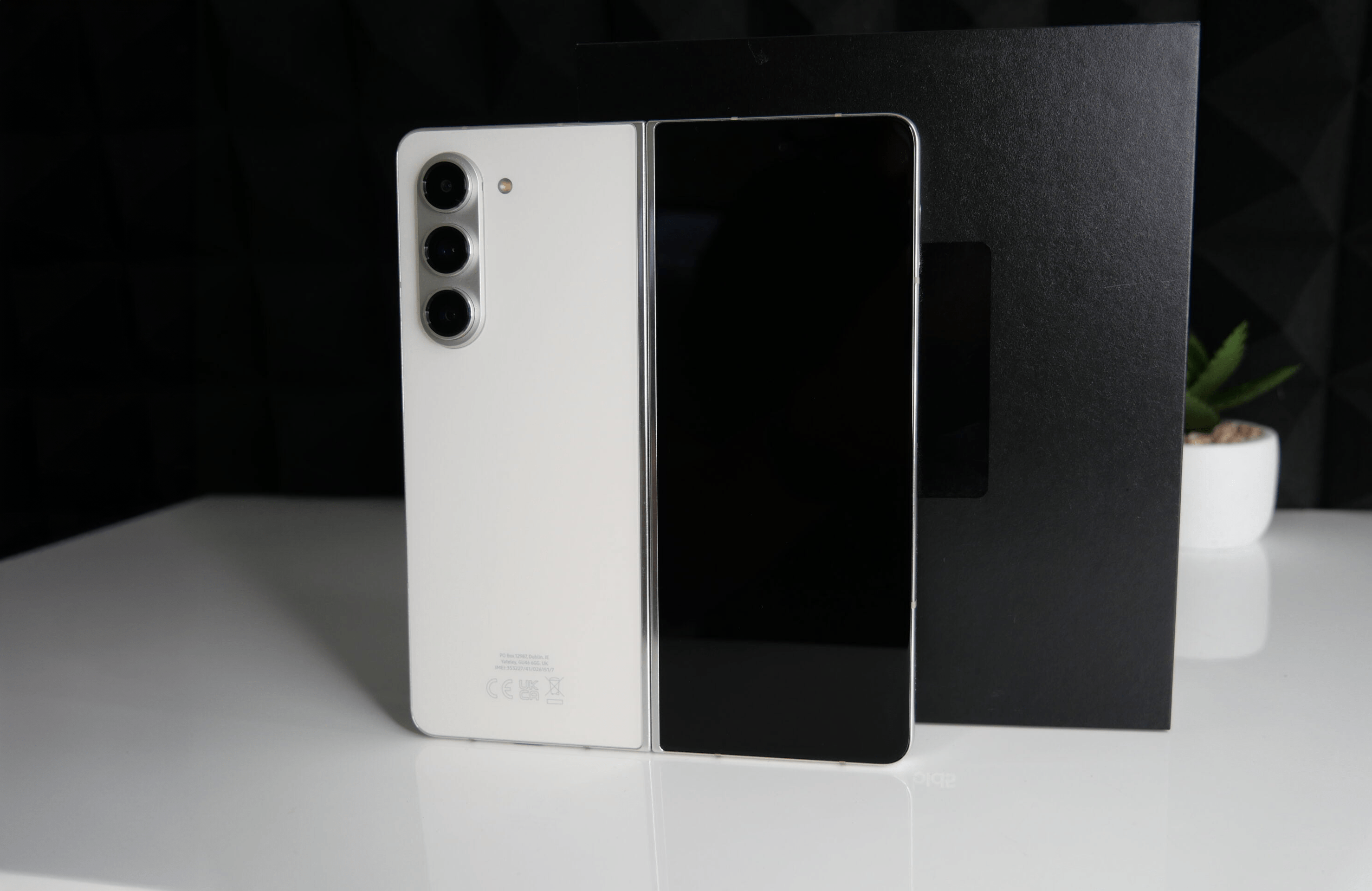 Biały składany smartfon z potrójnym aparatem, położony na białym stole przed czarnym tłem z geometrycznym wzorem.