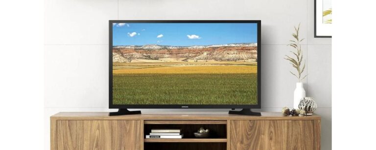 Telewizor stojący na drewnianej szafce z wazonem po prawej stronie. Na ekranie wyświetlany jest krajobraz z polami, wzgórzami i niebem.