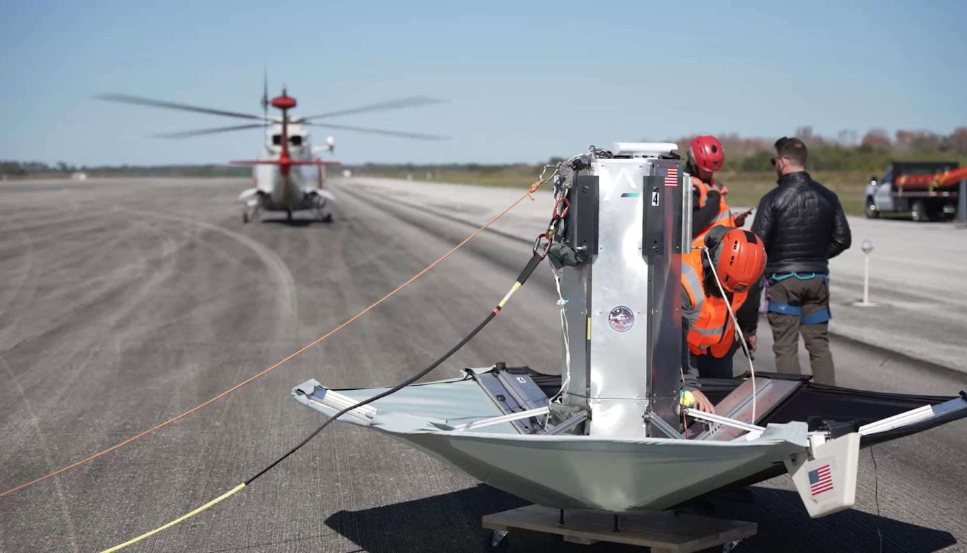 Osoby w kaskach i kamizelkach odblaskowych przy sprzęcie Sierra Space Ghost, w tle widoczny helikopter na pasie startowym.