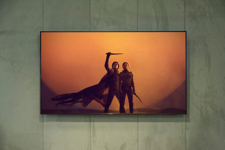 Dwie postacie w kostiumach bojowych stoją z bronią na tle pomarańczowego nieba, plakat filmowy zawieszony na ścianie.