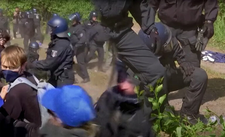 Funkcjonariusze policji w pełnym wyposażeniu interweniują podczas zamieszek, próbując obezwładnić protestujących w terenie zielonym.