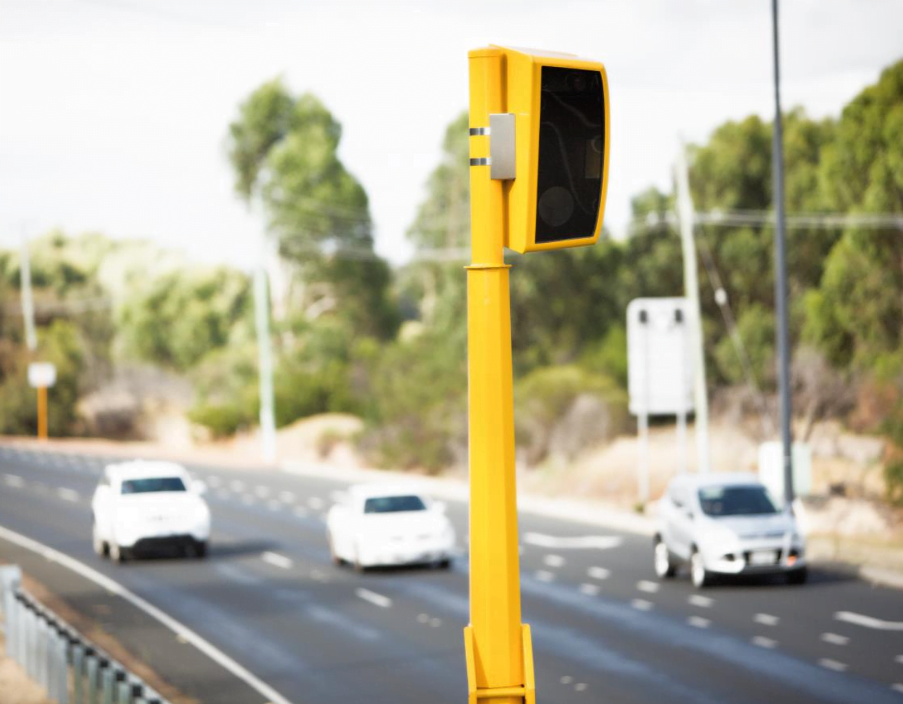 Żółty fotoradar na poboczu drogi z samochodami przejeżdżającymi w tle.
