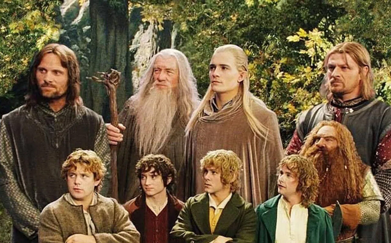 Uma foto do filme O Senhor dos Anéis. Um grupo de personagens de fantasia reunidos em um cenário de floresta, incluindo quatro hobbits, um homem com um cajado, um bruxo de barba branca, um elfo de cabelos claros, um homem de armadura e um anão de barba ruiva.