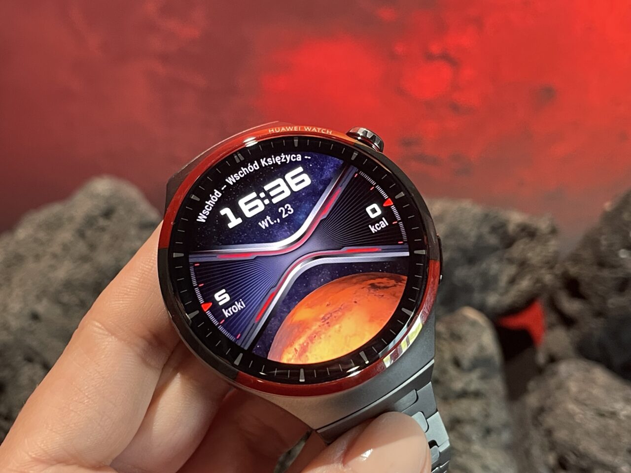 Zegarek Huawei na ręce osoby, z wyświetlaczem pokazującym godzinę i grafikę kosmiczną, w tle czerwone tło przypominające kosmos.