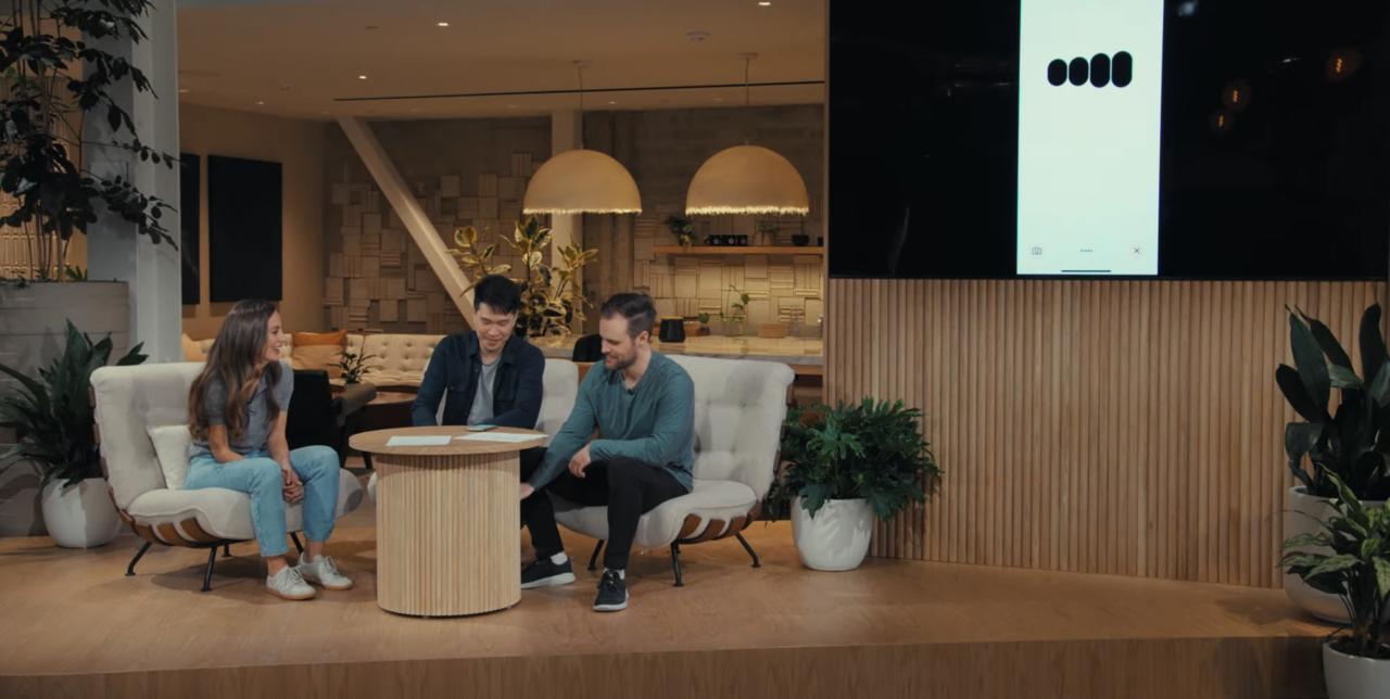 Trzy osoby siedzą w stylowej przestrzeni biurowej, dwie z nich oglądają ekran smartphone'a trzymanego przez mężczyznę.