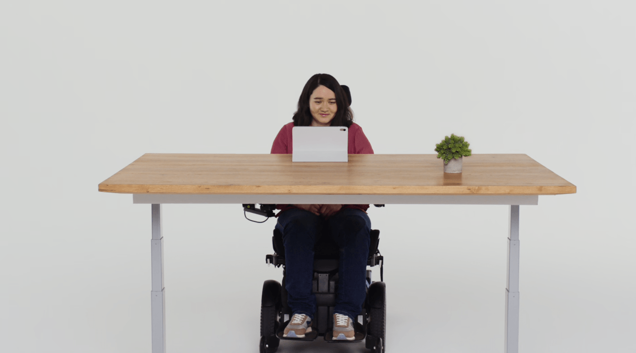 Osoba na wózku inwalidzkim siedzi przy drewnianym stole, patrząc na laptopa, z rośliną doniczkową obok.