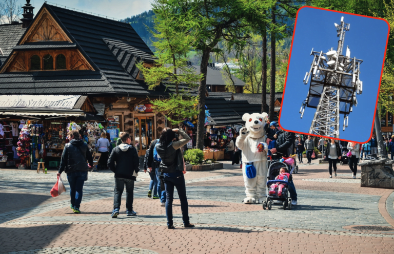 Tłum ludzi spacerujących po chodniku w górskiej miejscowości turystycznej, maskotka w przebraniu misia, drewniany budynek z pamiątkami, wstawione zdjęcie masztu telekomunikacyjnego.