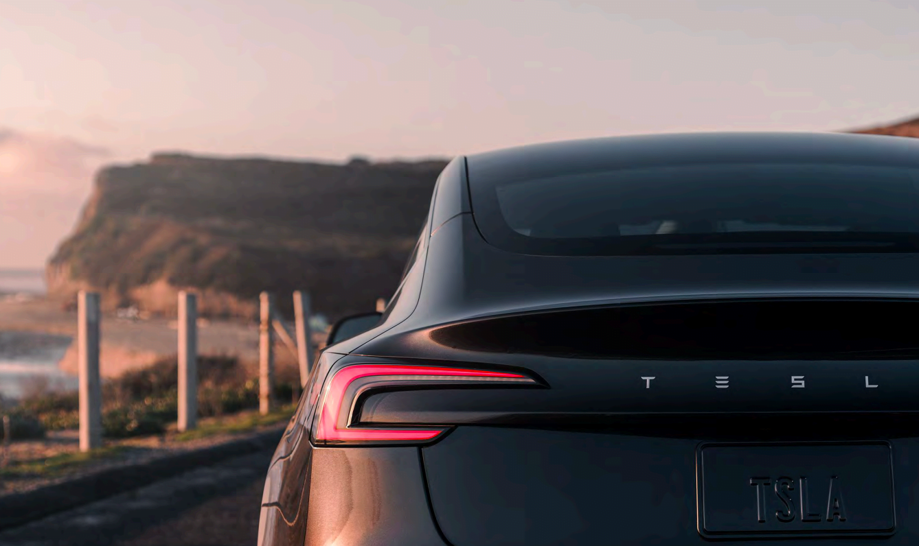 Tył czarnego samochodu Tesla z włączonymi światłami, na tle nadmorskiego krajobrazu.