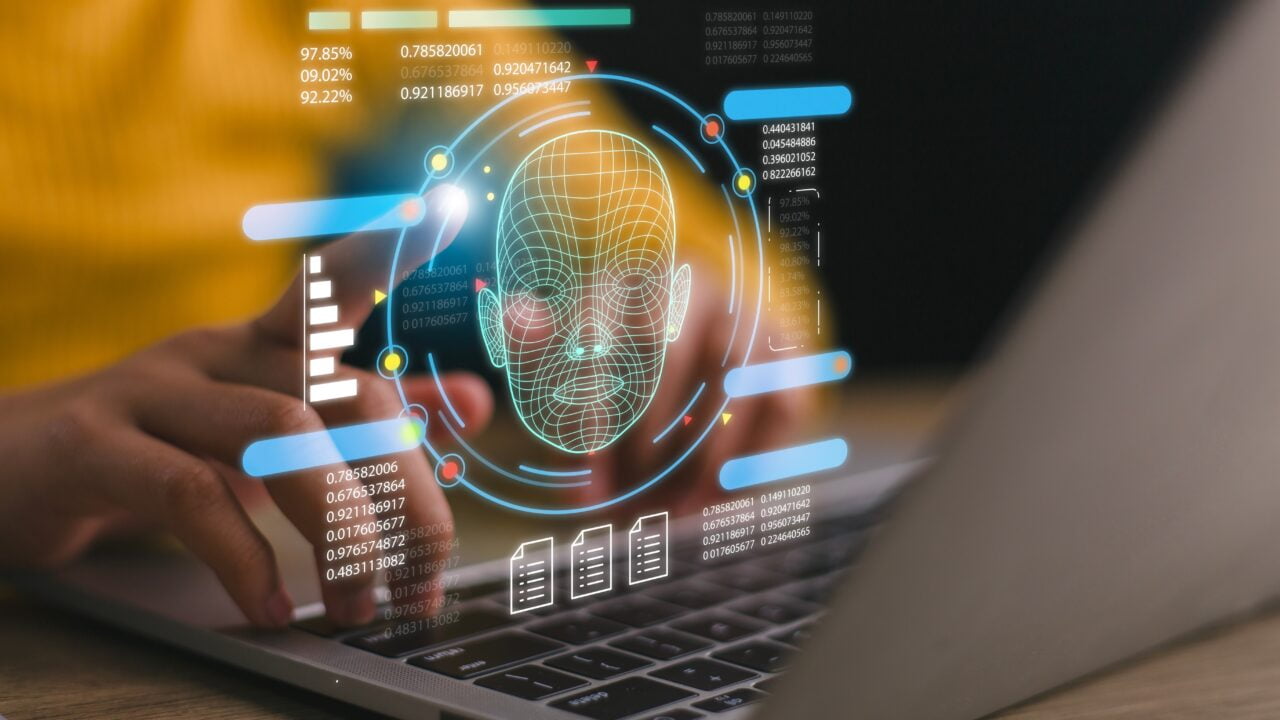 Osoba korzystająca z laptopa z wyświetlaną grafiką twarzy i danymi na ekranie, ilustrującą technologię rozpoznawania twarzy.