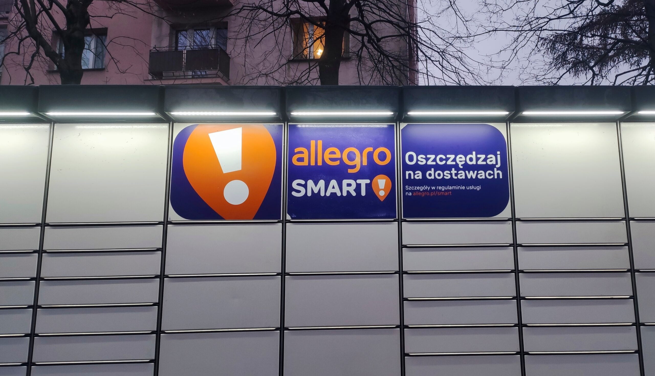 Tablica paczkomatu Allegro z logo "Allegro Smart!" i napisem "Oszczędzaj na dostawach".