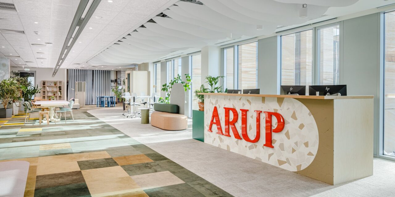 Nowoczesne biuro z recepcją oznaczoną dużymi czerwonymi literami "ARUP", ozdobioną jasną mozaiką. W tle widać jasne, przeszklone okna, rośliny doniczkowe, stoły i krzesła.