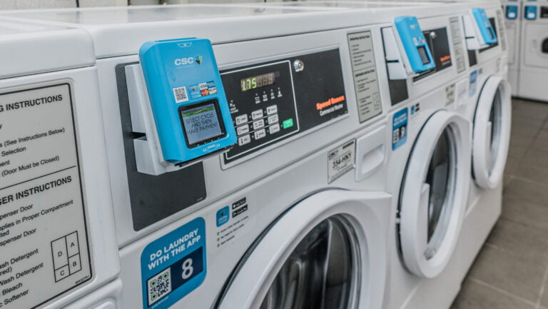 Kilka pralek w pralni samoobsługowej wyposażonych w urządzenia płatnicze CSC.