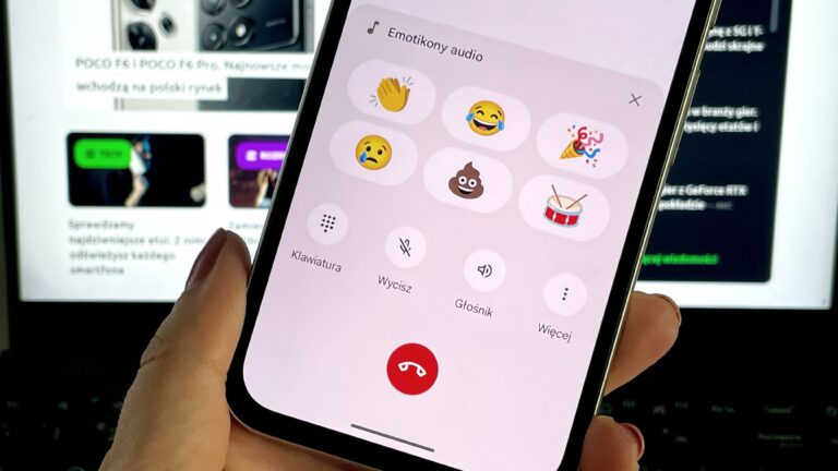 Smartfon trzymany w ręce z otwartą aplikacją "Emotikony audio" pokazującą różne emoji, w tle monitor komputera.