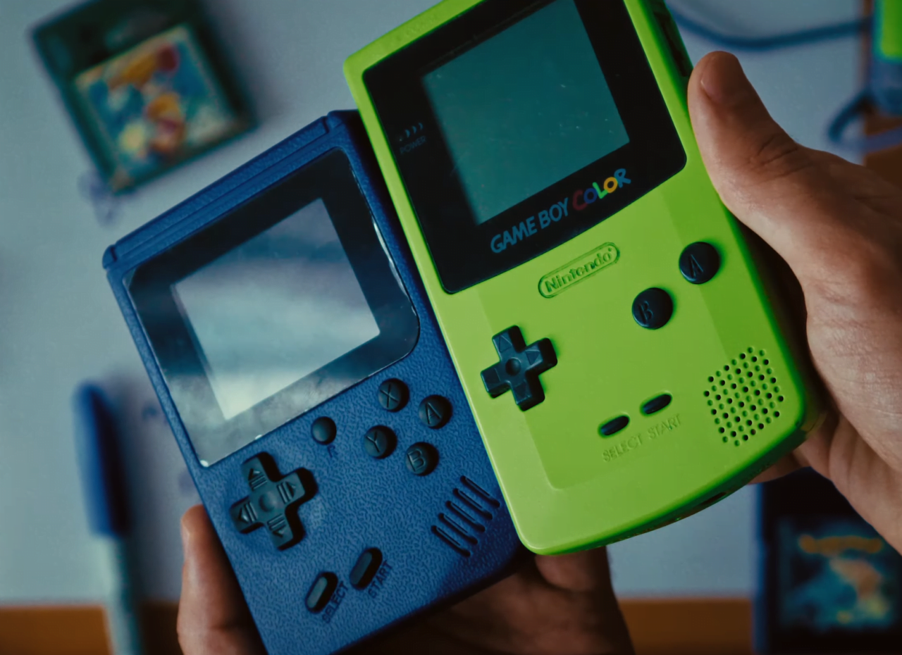 Dwie konsole Game Boy Color, jedna niebieska, druga zielona, trzymane w ludzkiej dłoni, z rozmytym tłem przedstawiającym opakowanie gry.