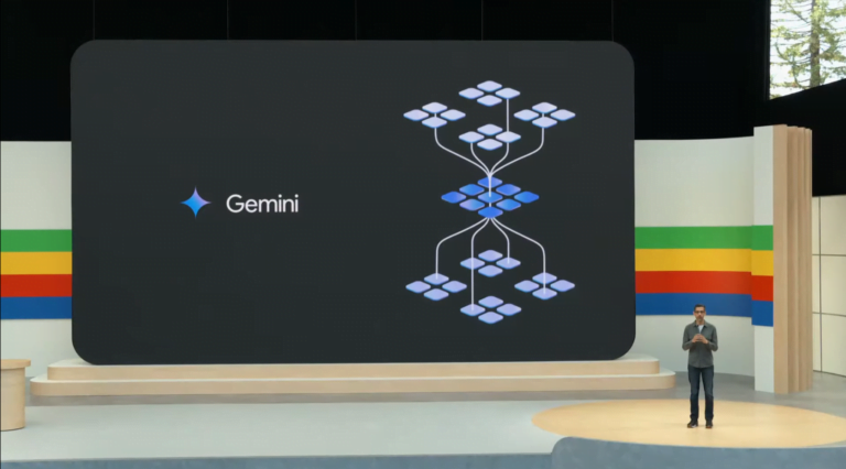 Mężczyzna na scenie prezentuje slajd z napisem "Gemini" oraz schematem blokowym na dużym ekranie w tle.