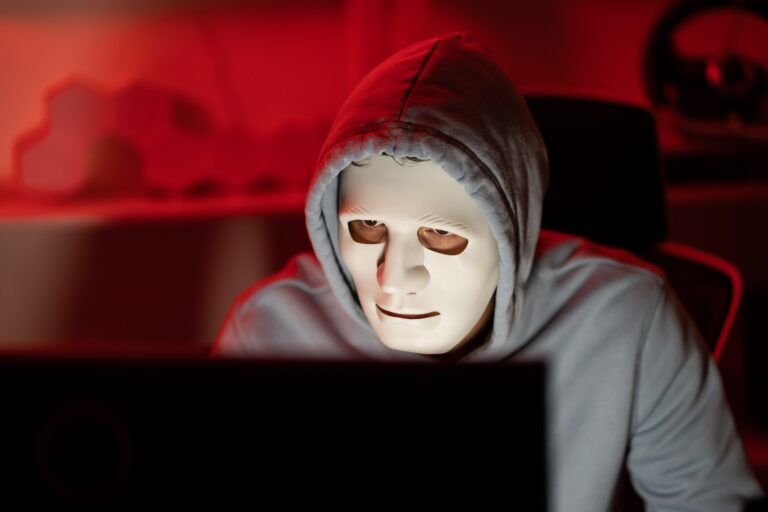 Osoba w masce i szarym kapturze siedzi przed ekranem komputera w czerwonym oświetleniu.