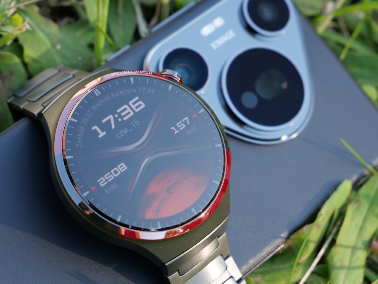 Smartwatch z metalową kopertą i bransoletą leżący na smartfonie z trzema aparatami, na tle trawy.