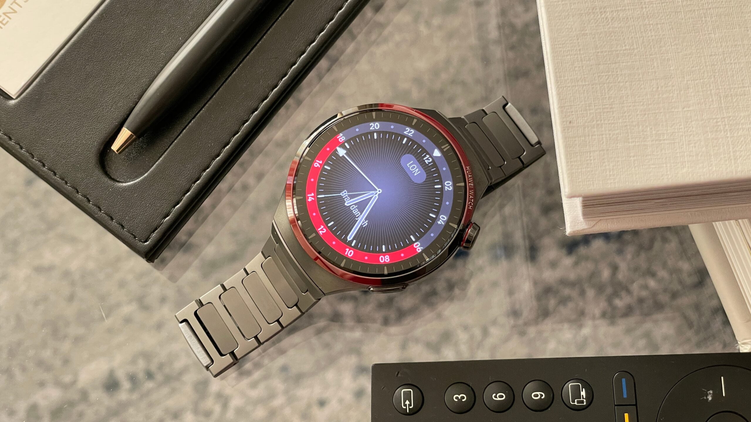 Smartwatch Huawei Watch z metalową bransoletą, czarnym skórzanym notesem, piórem i pilotem do telewizora na stole.