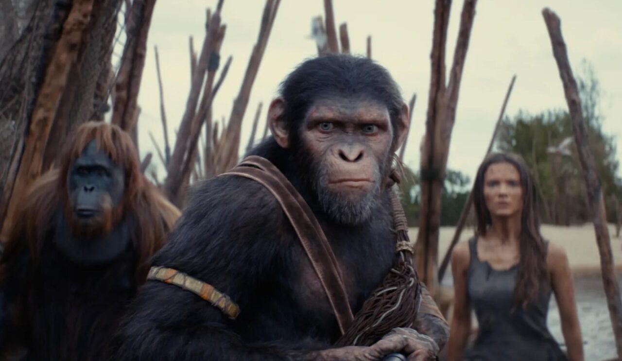 Scena z filmu z trzema postaciami, gdzie na pierwszym planie znajduje się postać przypominająca małpę w ludzkich ubraniach, na drugim planie widać kolejną małpę oraz kobietę w tle, stojących na piaszczystym terenie z drewnianymi konstrukcjami.