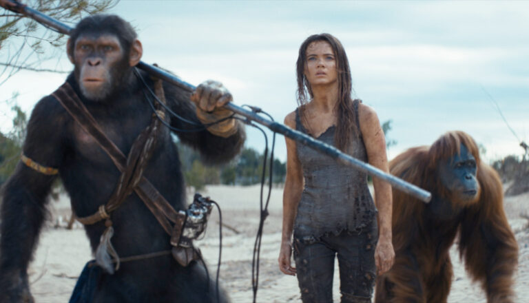 Kobieta i dwa małpy stoją na piaszczystym terenie z patykami w rękach.