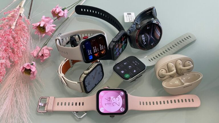 Różne modele smartwatchy i bezprzewodowe słuchawki na stole obok dekoracyjnych różowych kwiatów.