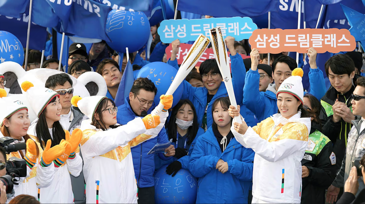 Samsung Galaxy Unpacked dwa dni przed olimpiadą. Ludzie ubrani w niebieskie i białe kurtki trzymają flagi i balony w trakcie sztafety olimpijskiej, dwóch uczestników wymienia się pochodniami olimpijskimi.